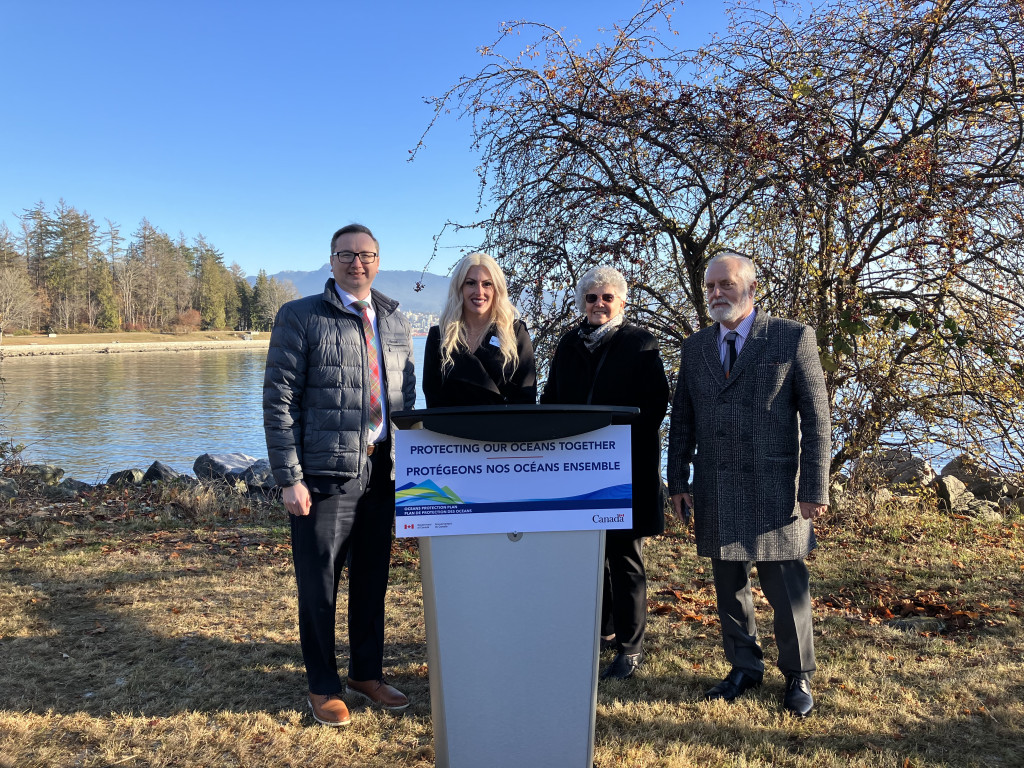 Four people stand behind a podium that is placed on grass next to a lake on a cold, dry winter day