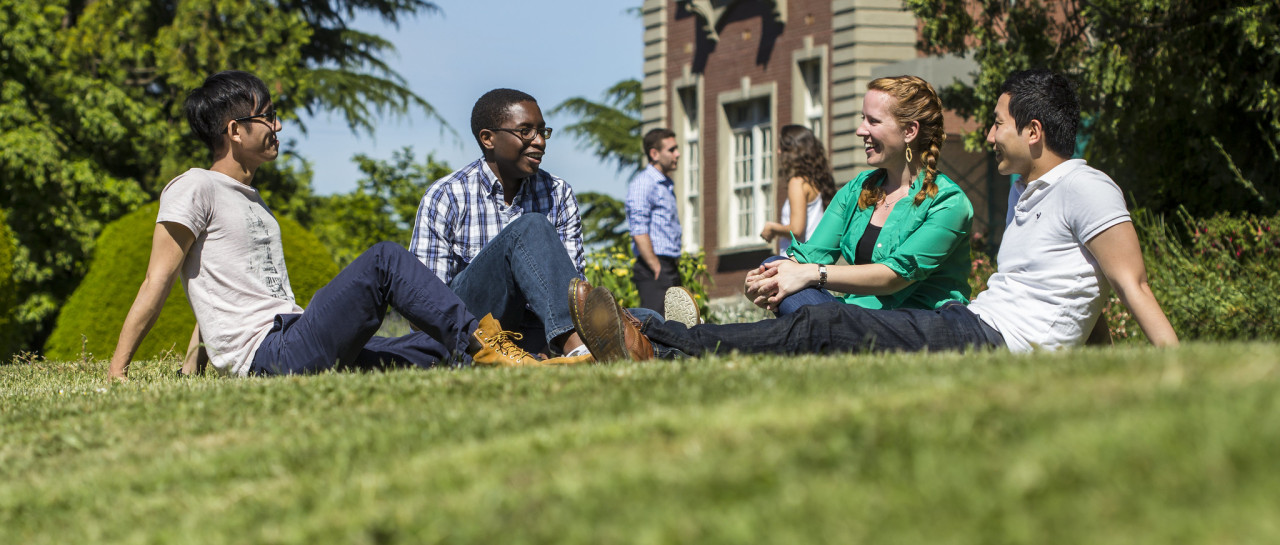 A group of 5 students sitting on the grass. They are talking and laughing. 