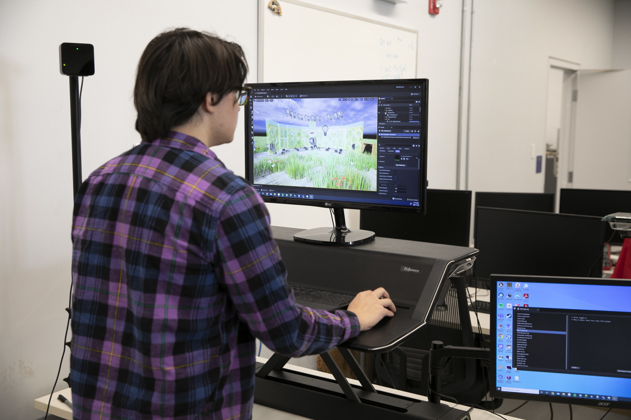 A man in a purple plaid shirt stands in front of a computer screen with a virtual reality design.